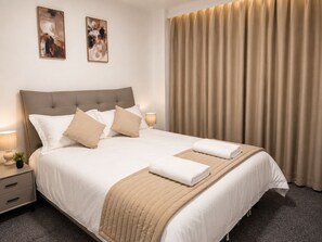 A hotel room with a large bed, two bedside tables, a nightstand, a lamp, a window with curtains, and two wall-mounted paintings.