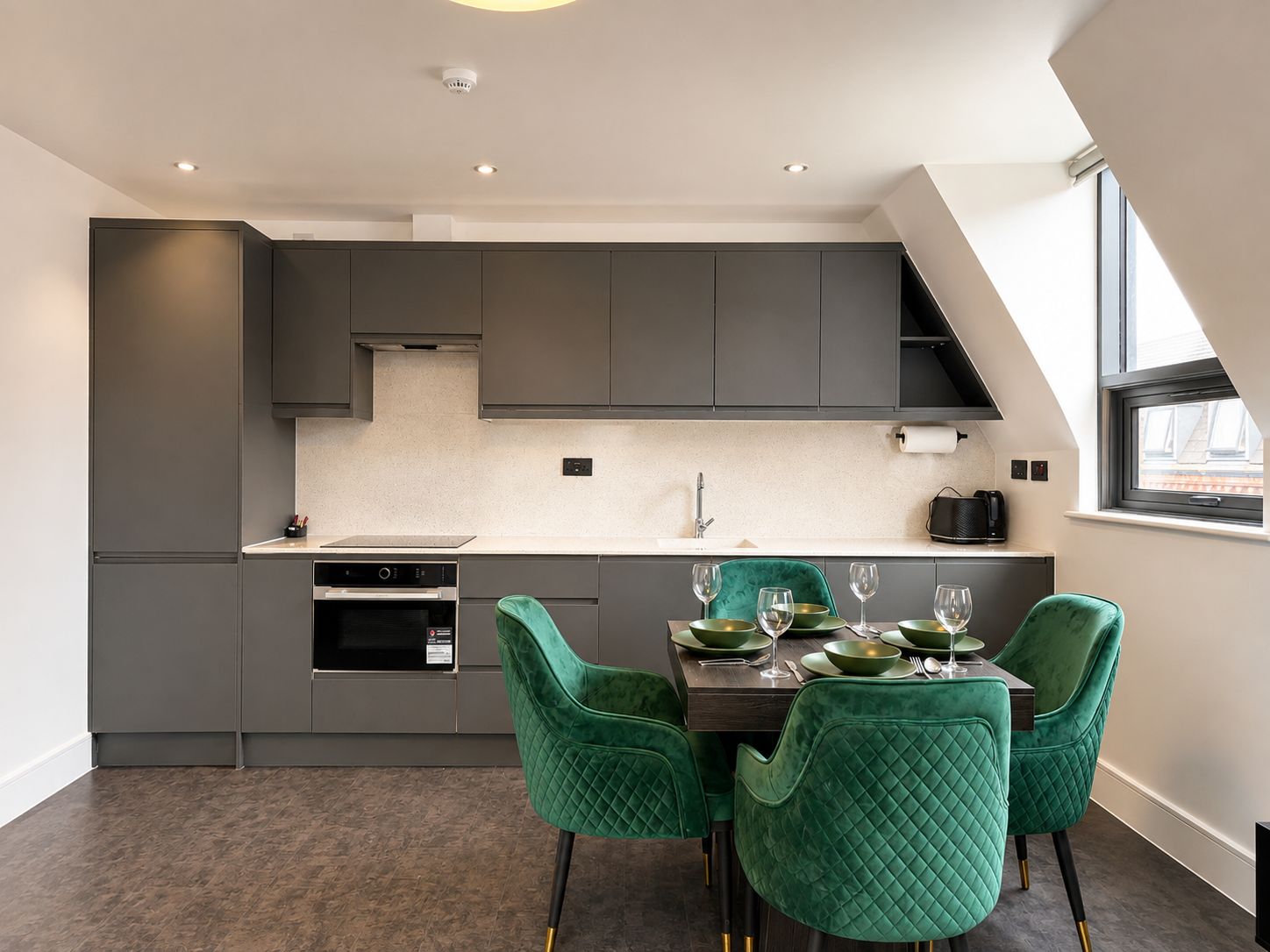 A modern kitchen with dark cabinetry, a central island, and a dining area with green upholstered chairs.