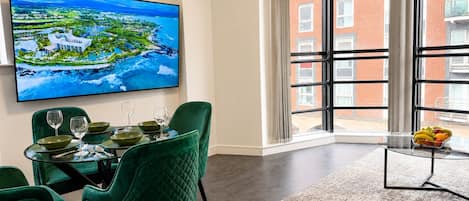 A dining area with a table set for four, a large window with a view of a red brick building, and a painting of a coastal landscape.
