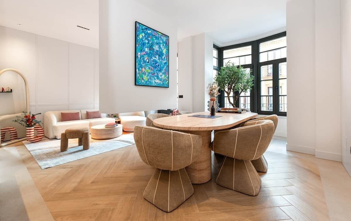 Een moderne woonkamer met een ronde eettafel, beige banken en een groot raam met uitzicht op gebouwen.