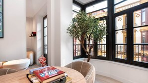 Een kamer met een ronde tafel, boeken, een kamerplant en grote ramen met uitzicht op gebouwen.