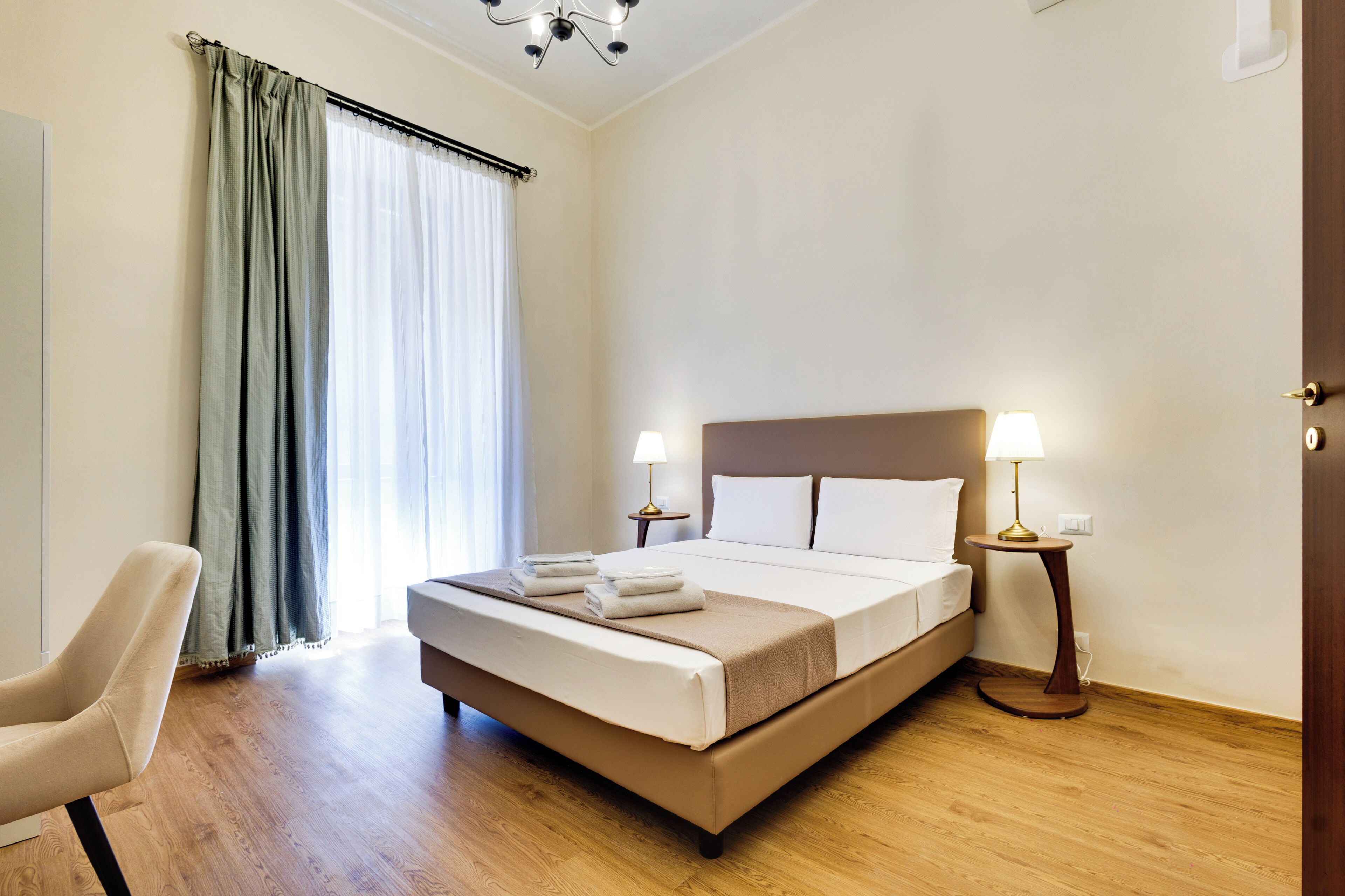 A hotel room with a bed, two bedside lamps, a chair, and a window with curtains.