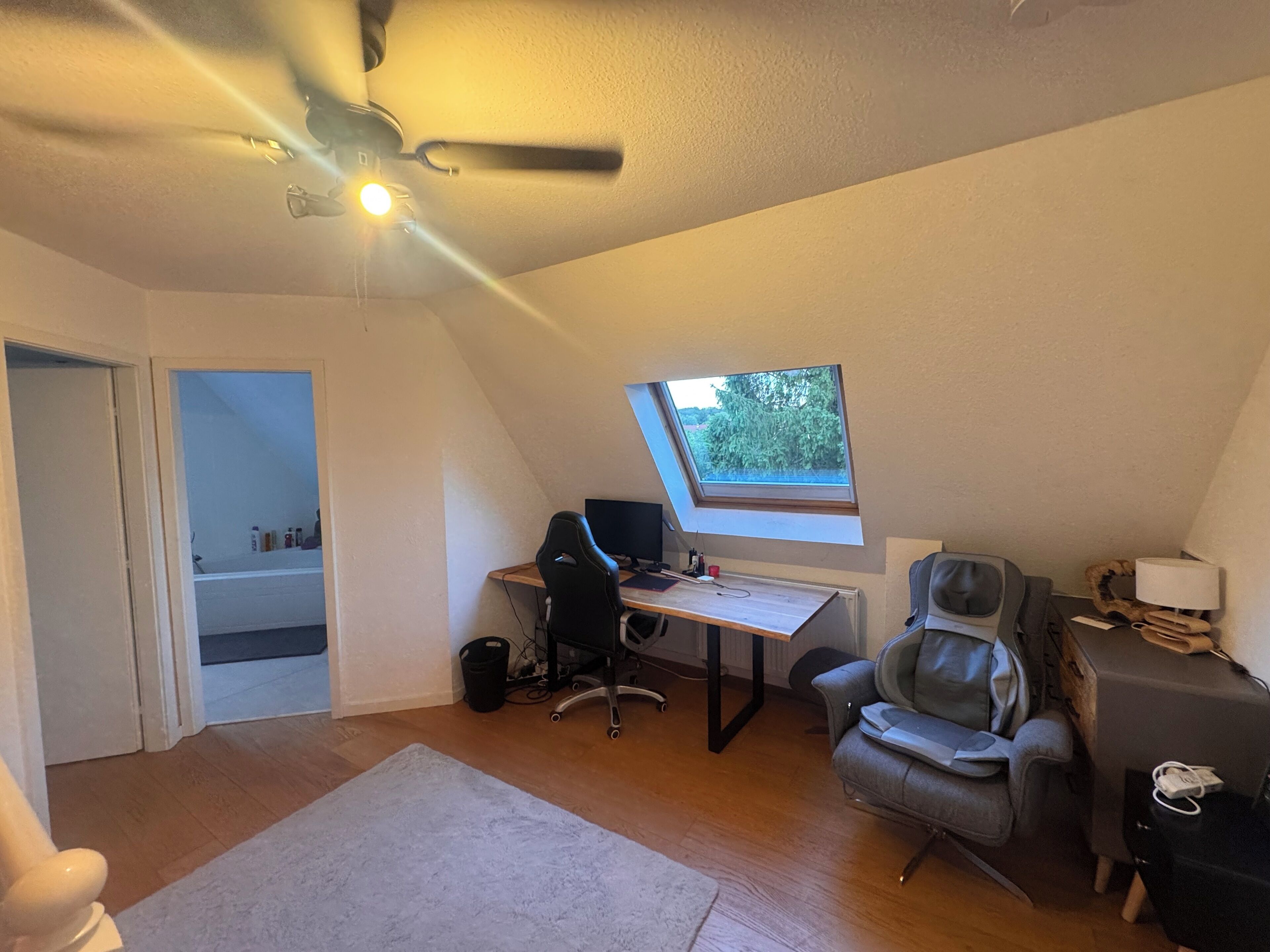 Una habitación con techo inclinado, un escritorio con computadora, una silla gamer, un sofá y una ventana con vista a follaje.