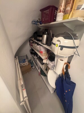 A narrow storage area with shelves containing various items including a vacuum cleaner, a blue umbrella, and some packaged goods.