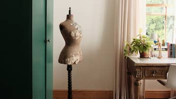 A room with a wooden floor, a green cabinet, a mannequin, a desk with a lamp, a potted plant, and a window with curtains.