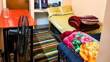 A compact room with a bed, a table, a fan, and a colorful rug.