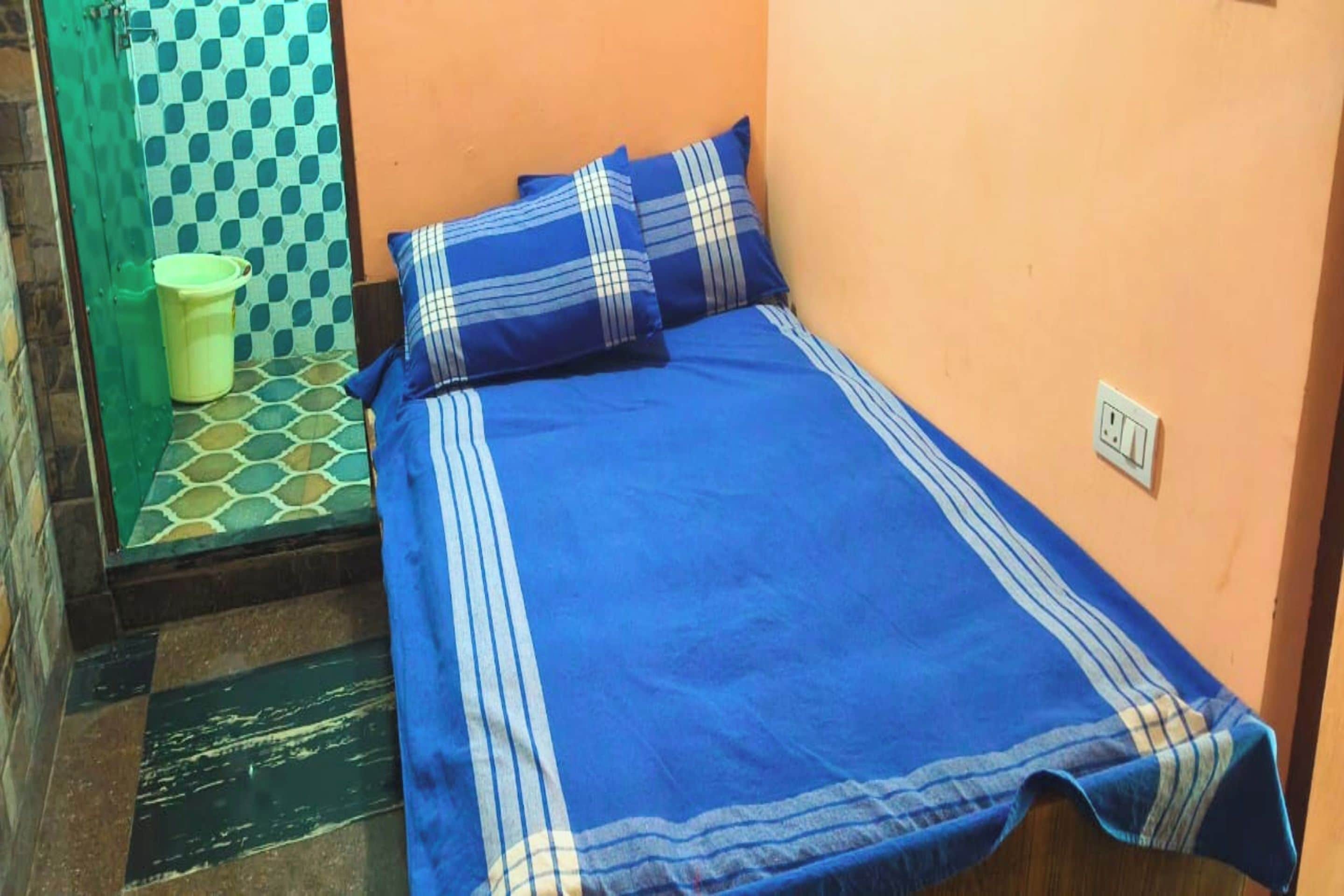 Habitación pequeña con una cama individual, colcha azul y almohadas a juego, un balde verde y un papel tapiz estampado.