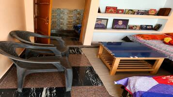 A room with a bed, a small table, two chairs, and a shelf with framed photos.