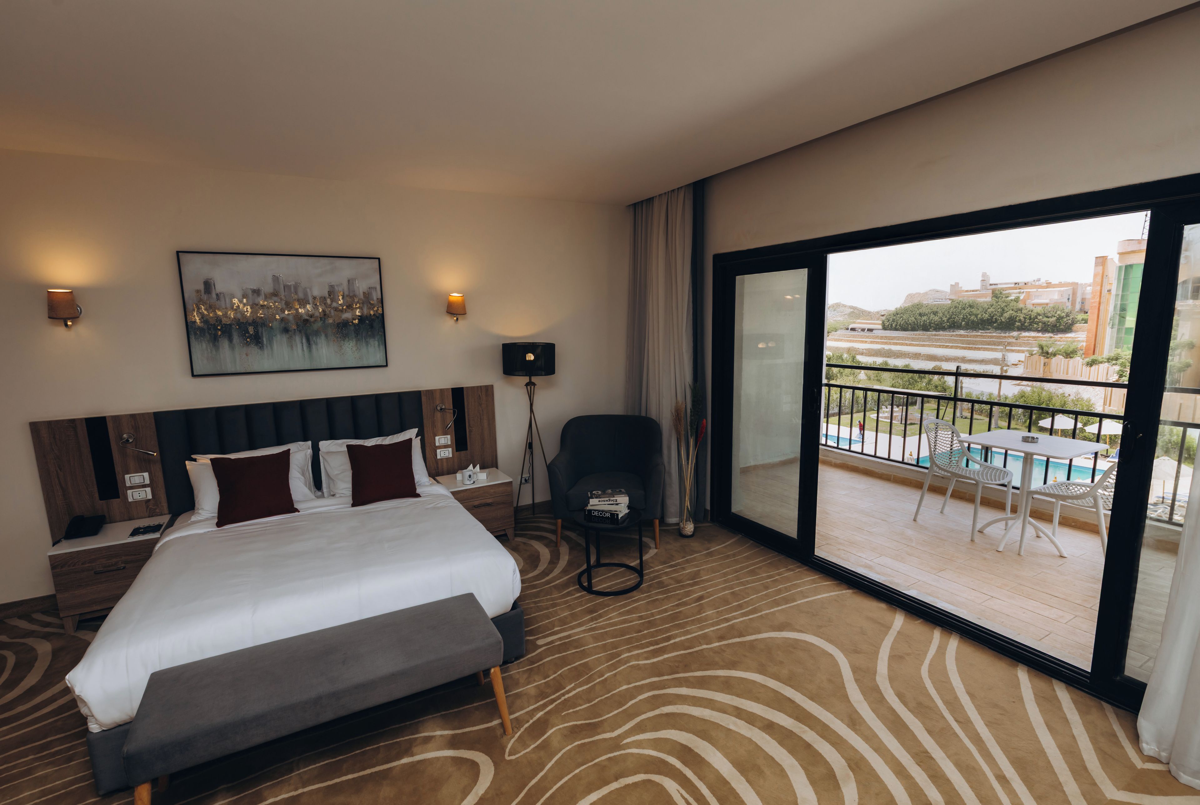 A hotel room with a bed, bedside tables, a chair, a painting, and a view of a swimming pool and buildings.