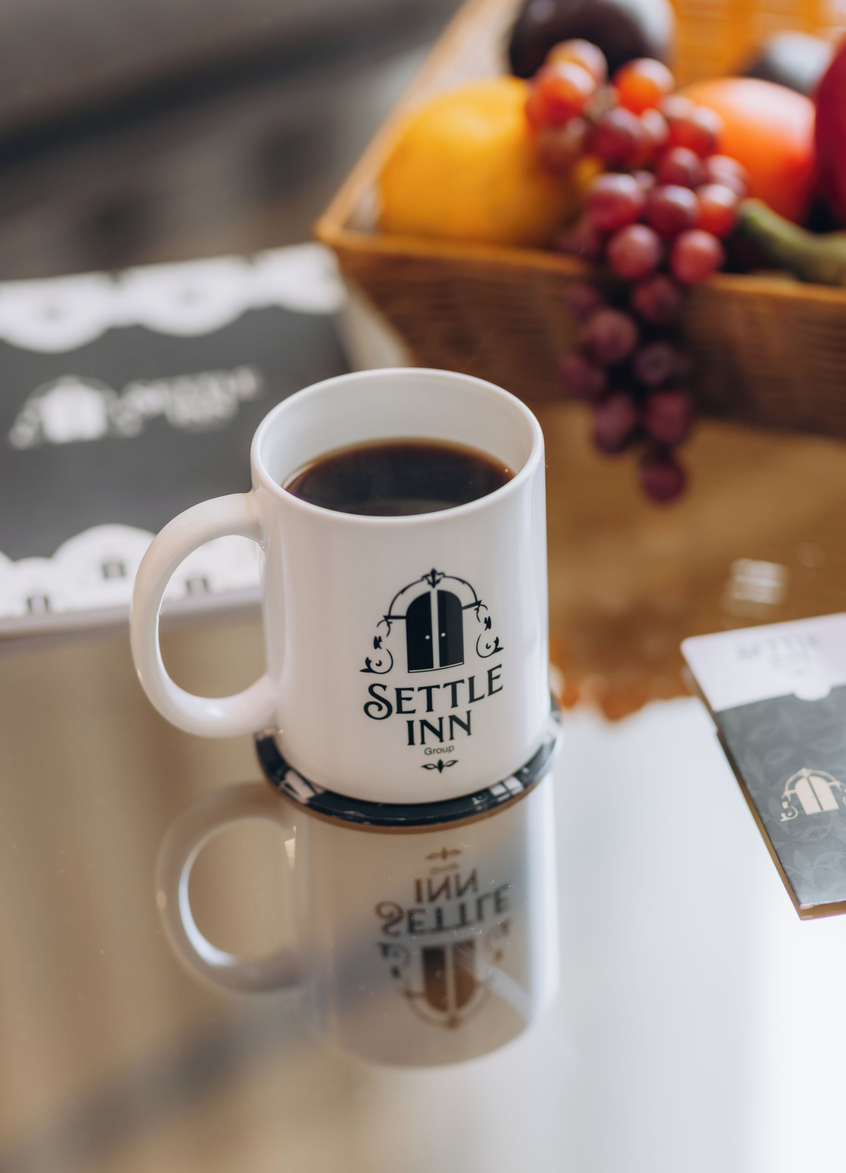 A white coffee mug with 'SETTLE INN Group' logo on a coaster, a basket of fruits, and a card with a bell icon.