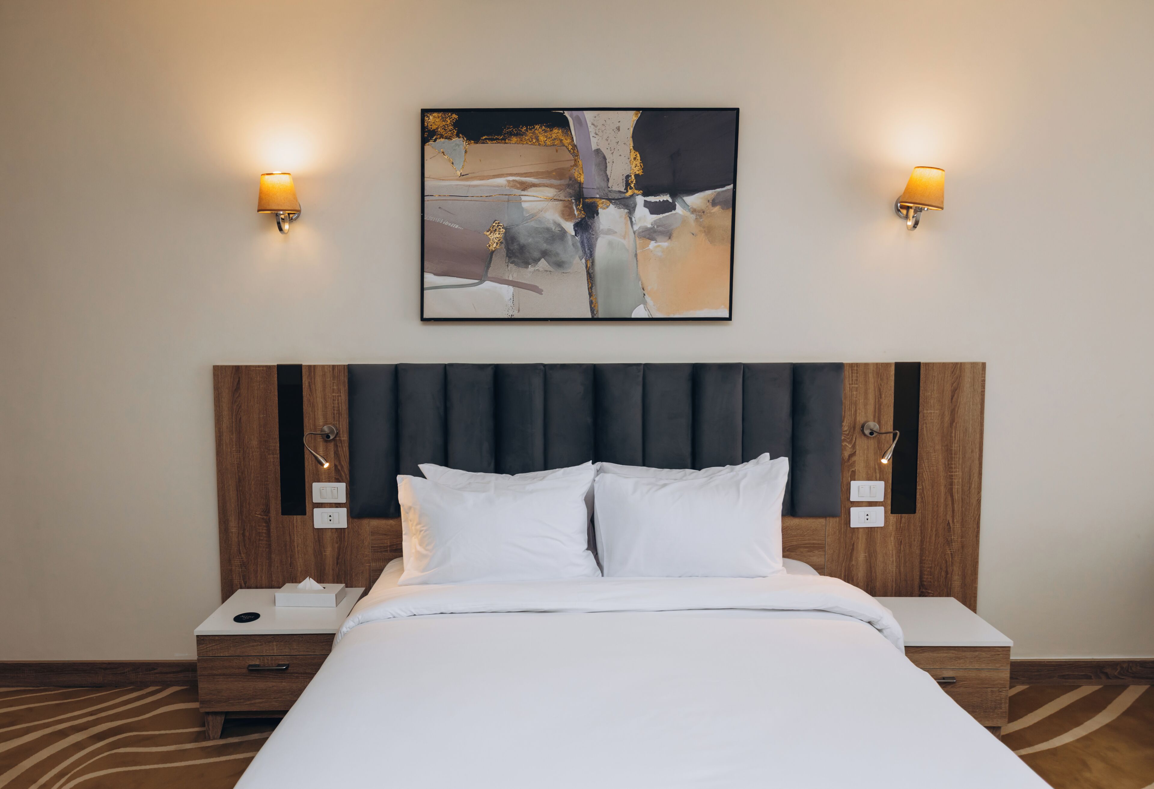 Una habitación de hotel con una cama grande, dos mesitas de noche, un rabillo de la cama, un cuadro abstracto colgado en la pared y dos lámparas de pared.