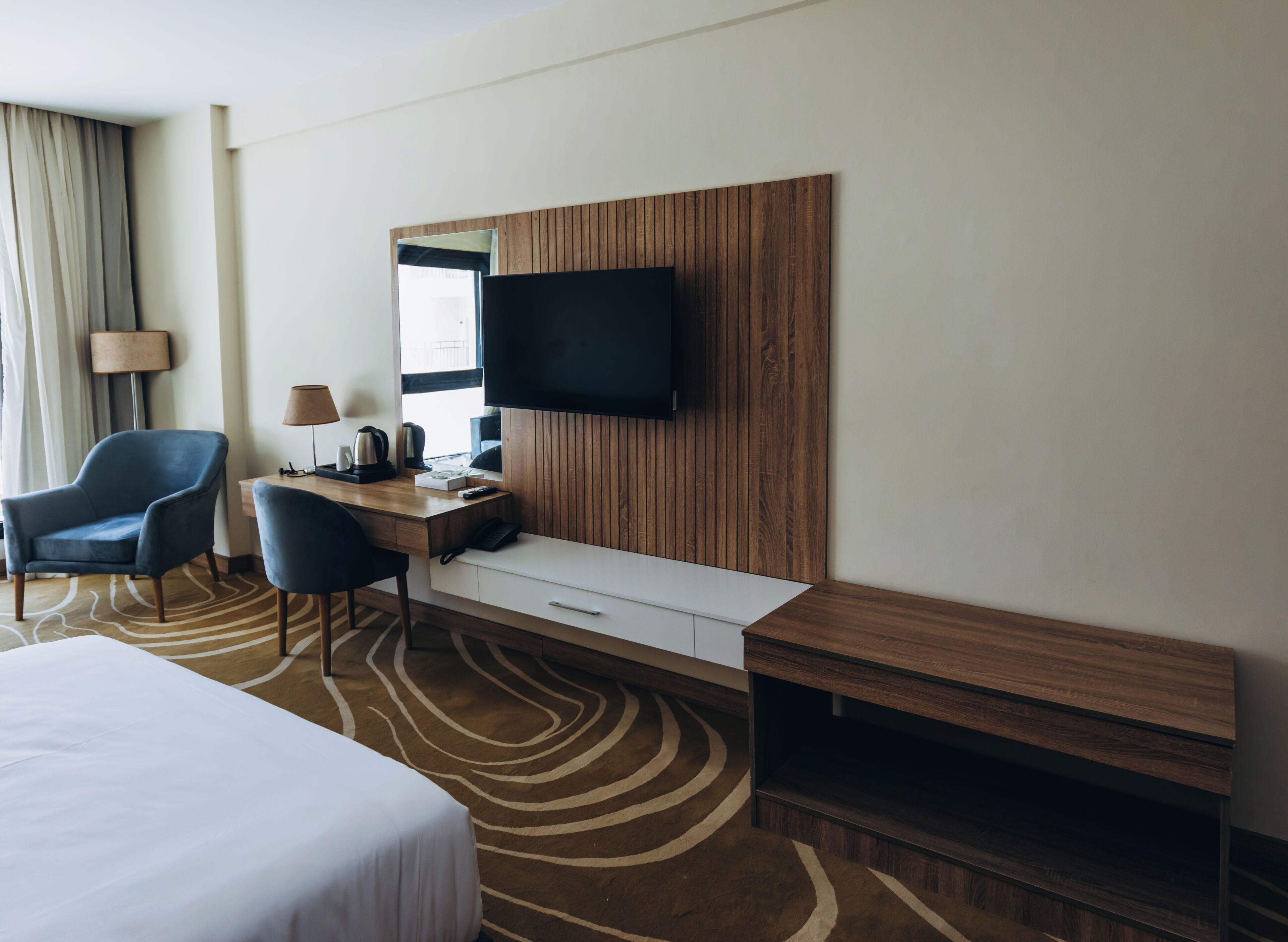 A hotel room with a bed, a desk with a chair, a television, and a window with curtains.