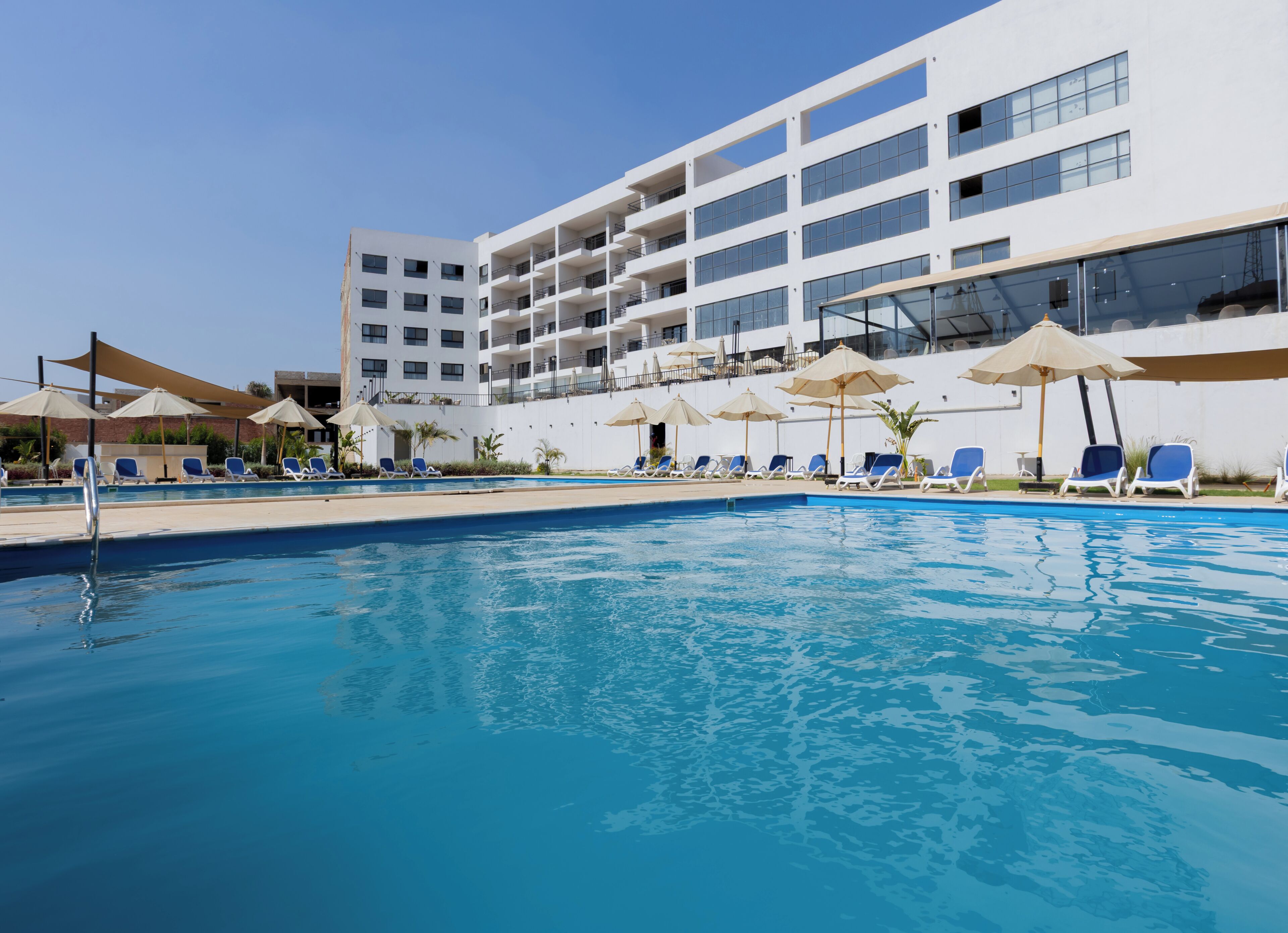 Une piscine entourée de chaises longues et de parasols, le tout bordé d’un immeuble moderne doté de nombreux balcons.