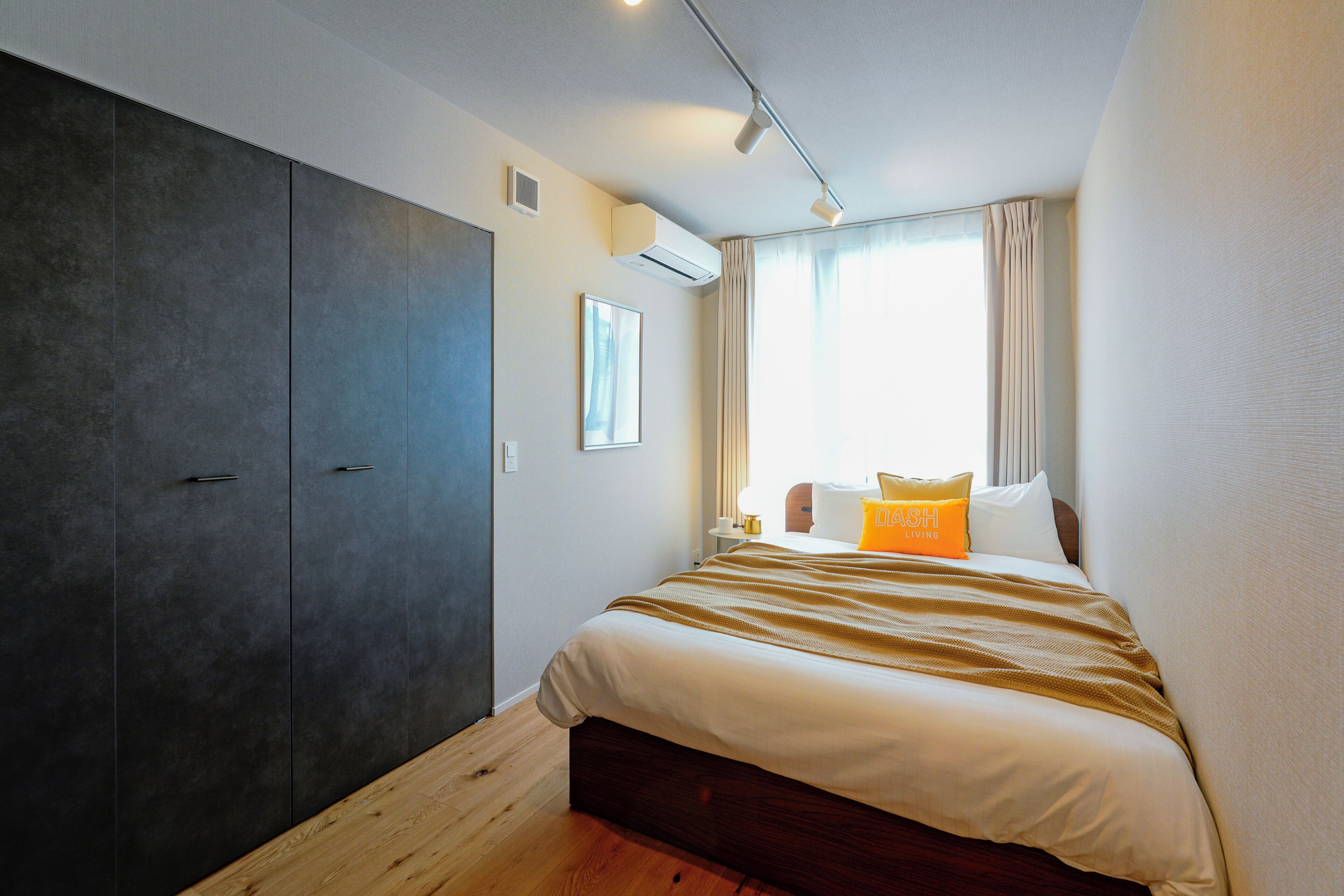A bedroom with a large wardrobe, a bed with a striped blanket, a bedside table with a lamp, and a window with curtains.
