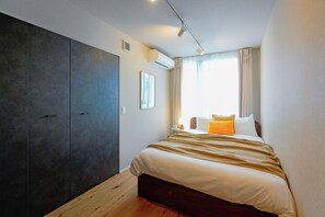 A bedroom with a large wardrobe, a bed with a striped blanket, a bedside table with a lamp, and a window with curtains. - Dash Living Nezu (Bunkyo-ku)