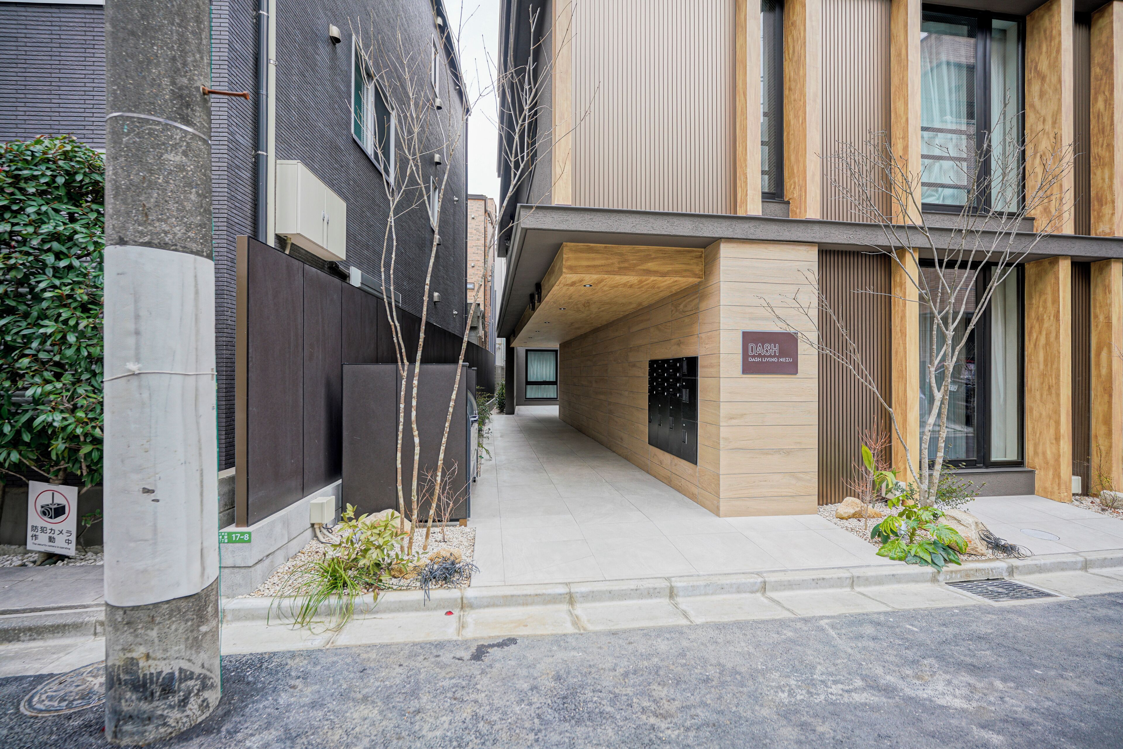 Un edificio moderno con fachada de madera, un cartel que indica 'DASH LIVING NEZU' y un estrecho callejón con árboles y un pequeño jardín.