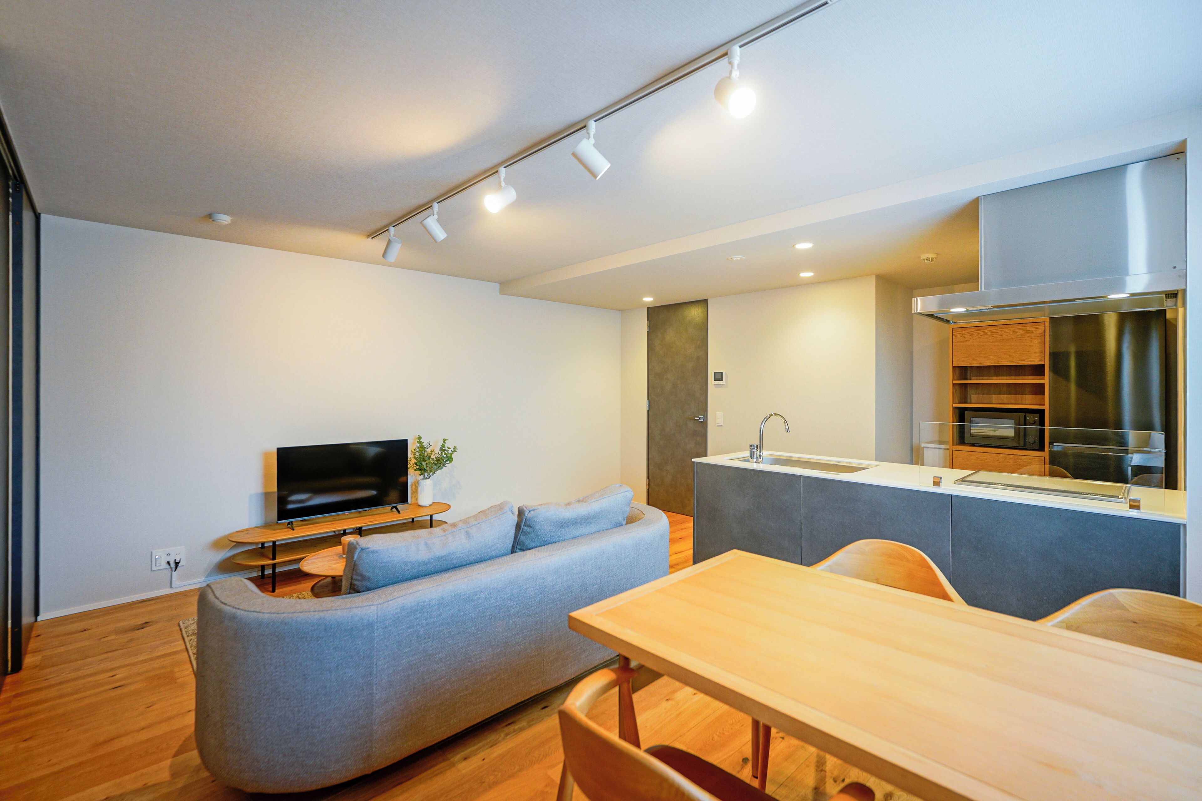 A modern living room with a grey sofa, a wooden dining table, and a kitchen area with a stainless steel range hood.