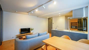 A modern living room with a grey sofa, a wooden dining table, and a kitchen area with a stainless steel range hood.