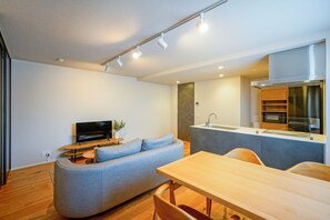 A modern living room with a grey sofa, a wooden dining table, and a kitchen area with a stainless steel range hood. - Dash Living Nezu (Bunkyo-ku)