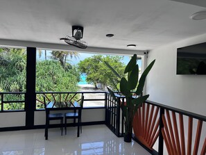 A balcony with a view of a beach and palm trees, a ceiling fan, and a potted plant.
