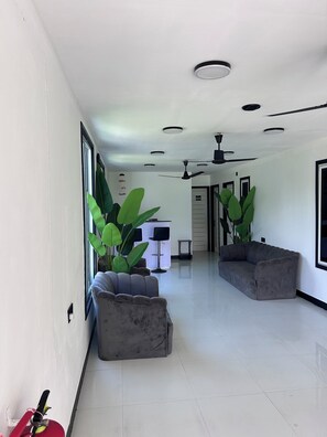 A spacious hallway with a grey sofa, large green plants, a ceiling fan, and recessed lighting.