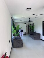Un pasillo amplio con un sofá gris, plantas verdes grandes, un ventilador de techo y luces empotradas.