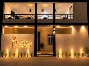 A two-story hotel with a balcony on the upper floor, a sign reading 'ALYRAA SUNSET,' and potted plants along the entrance.
