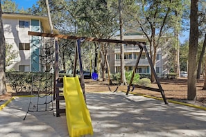 A playground with swings and a slide, surrounded by trees and buildings. - Fiddler's Cove - 2 Pool -Tennis - Hot Tub - Gym - Walk to the Beach! (Hilton Head Island)