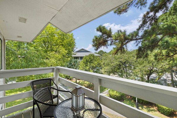 Uma varanda com mesa e cadeiras, vista para um jardim com árvores e um prédio ao fundo.