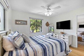 Quarto com cama, ventoinha no teto, televisão e janela com vista para árvores.