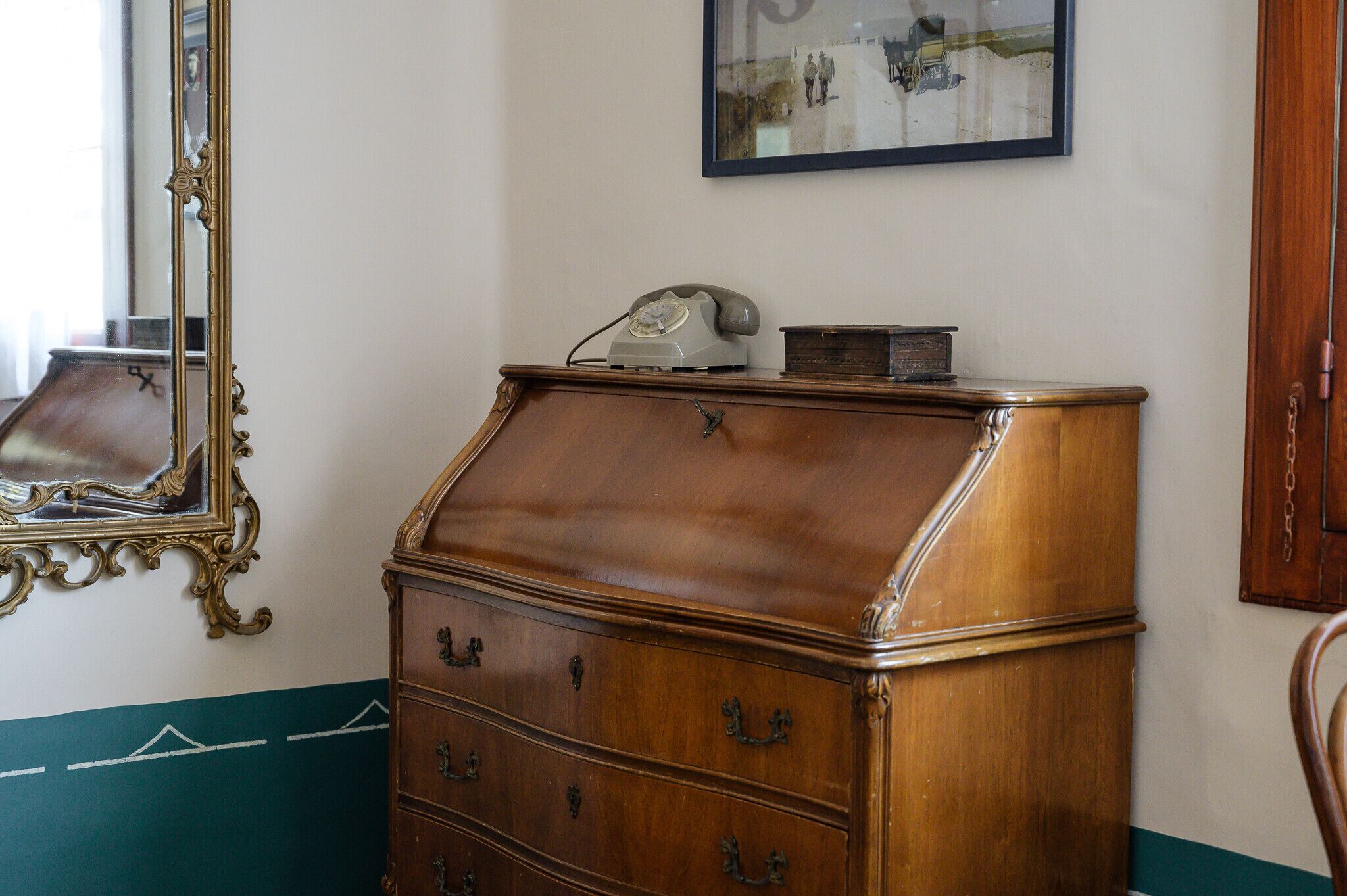A wooden chest of drawers with a telephone on top, a mirror with ornate frame, and a framed picture on the wall.