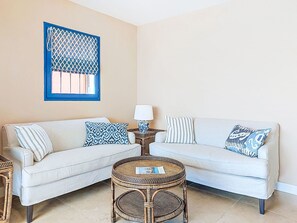 A living room with a beige wall, a blue-framed window, a white sofa with patterned cushions, a round wicker table, and a floor lamp. - Village de Silhouette by Interhome (Grimaud)