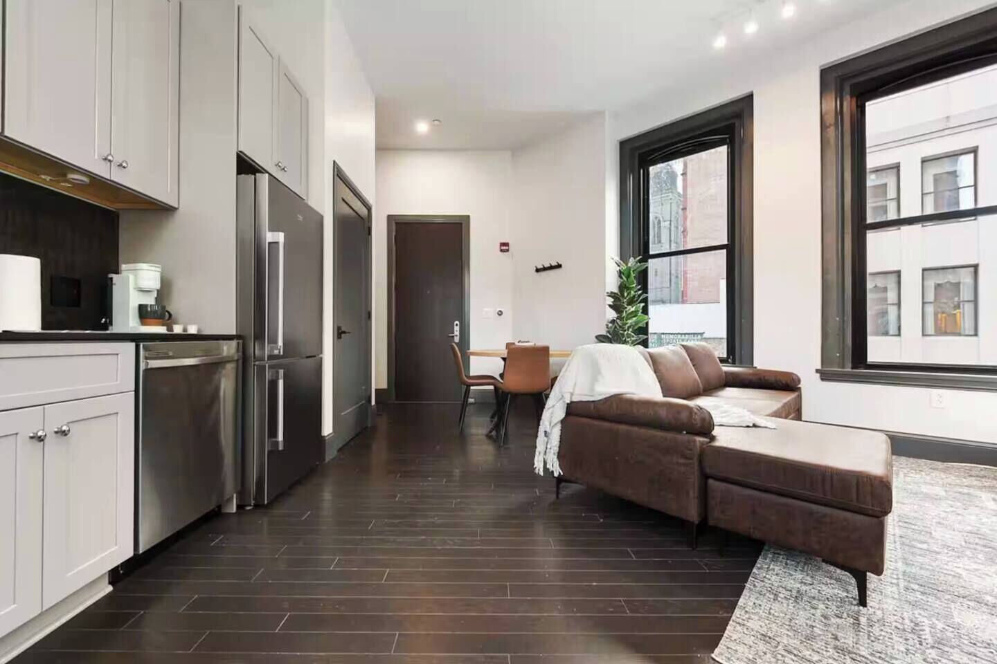 A modern kitchen with stainless steel appliances, a dining area with a brown leather couch, and large windows providing natural light.
