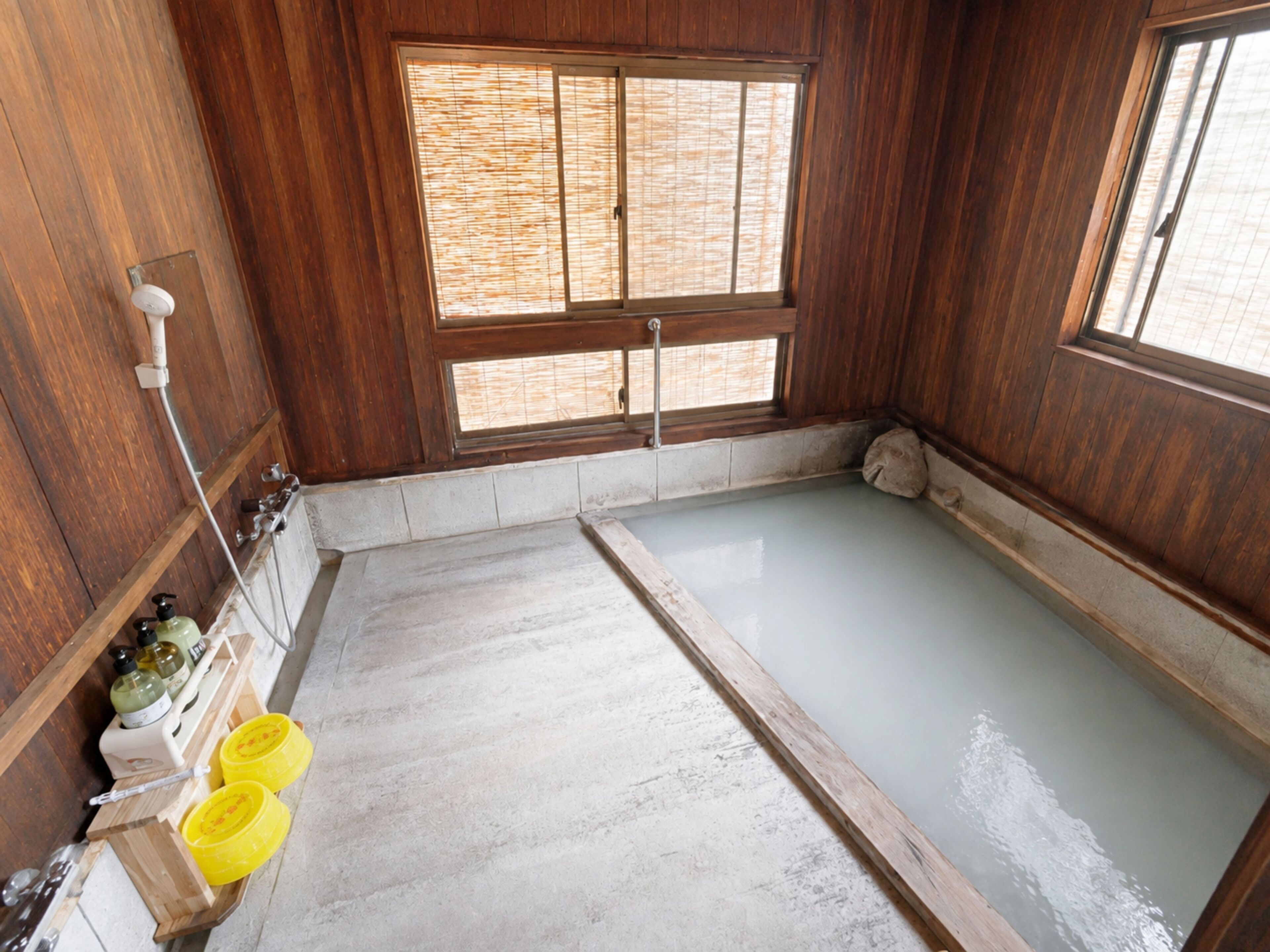 A room with a wooden floor, a window with bamboo blinds, a bathtub, and a shelf with toiletries.