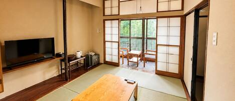 Ein traditioneller japanischer Raum mit Tatami-Boden, einem Fernseher, einem Tisch und Schiebetüren, die auf einen Balkon führen.