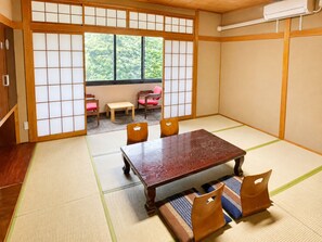 A tatami mat room with a wooden table and chairs, a sliding door leading to a balcony, and a view of greenery outside.