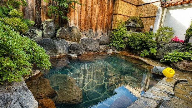 A clear water feature surrounded by rocks and greenery, with a wooden fence and a small building in the background.