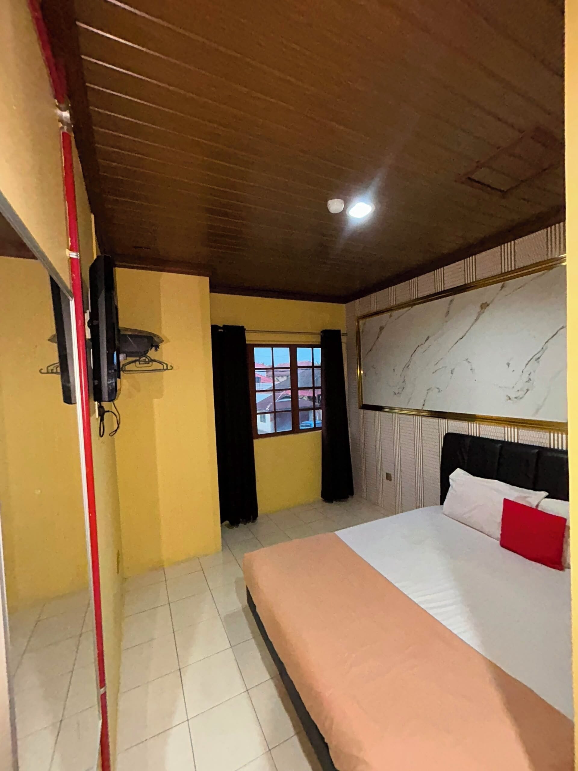 Una camera d'albergo con un letto, una finestra con tende, una televisione a muro e un lampadario al soffitto.