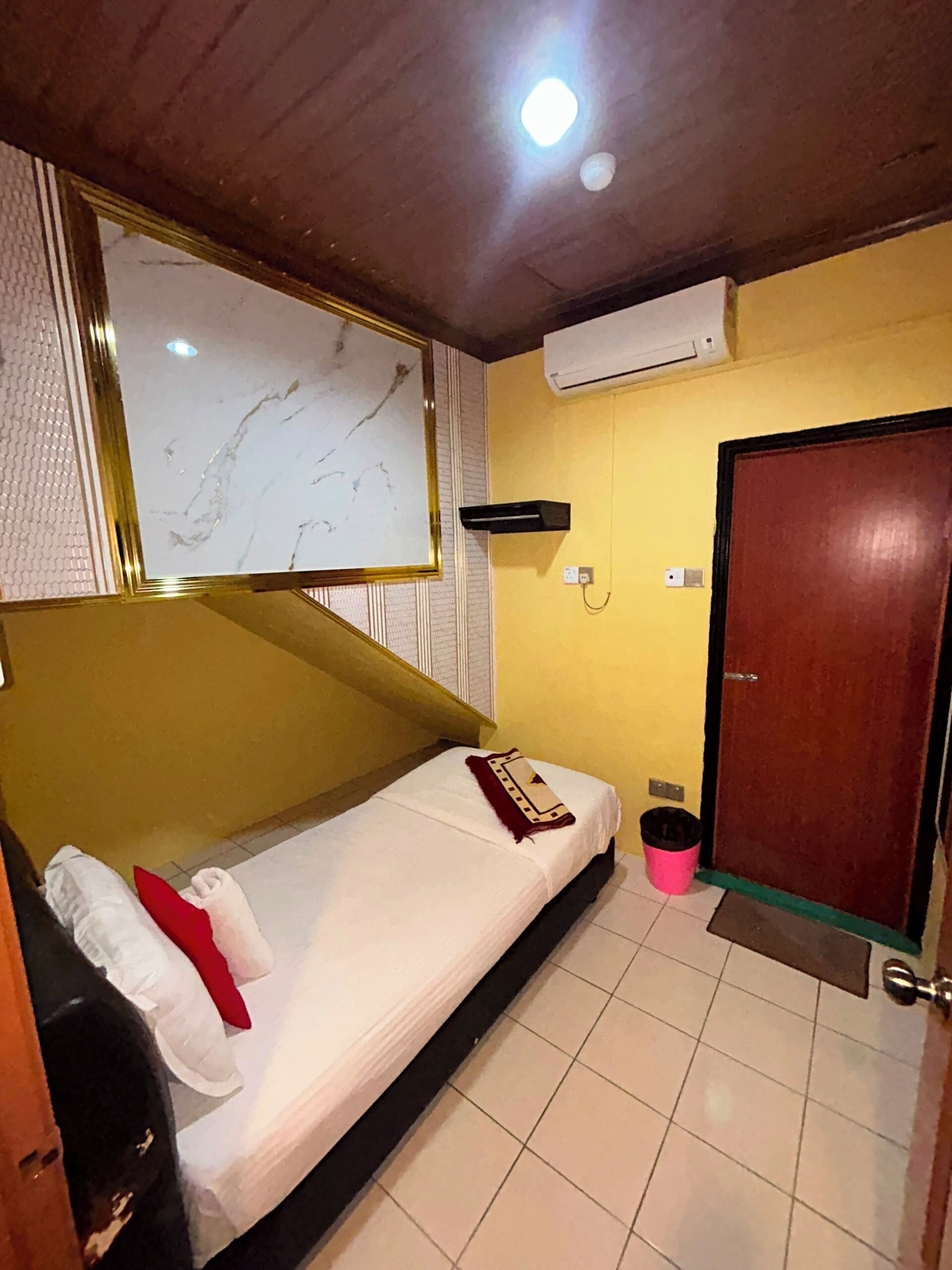 A single bed with a white mattress, a red pillow, and a beige blanket. There is a wall-mounted air conditioning unit, a wooden door, and a small trash bin.