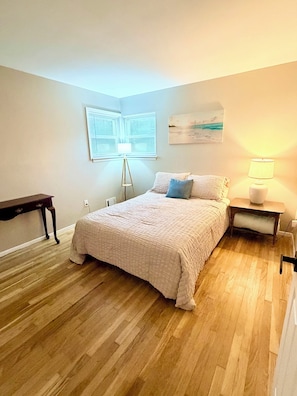 A bedroom with a bed, bedside tables, a lamp, a window with blinds, and a painting on the wall.