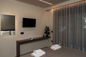 A modern hotel room with a flat-screen TV mounted on the wall, a wooden console table with a plant and candles, a large window with curtains, and two neatly folded towels on the floor.
