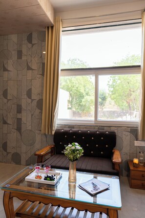 A modern living room with a glass-top coffee table, a dark brown sofa, a wooden side table, and a window with curtains. - ELIVAAS Signature Hiara Rudraksha 6BHK  (Udaipur)