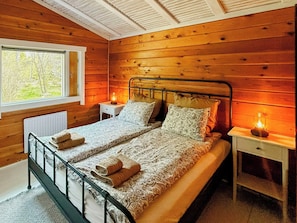 Ein Doppelbett mit gemusterten Bettbezügen und zwei Kissen, ein Nachttisch mit Lampe sowie ein Fenster mit Blick auf Bäume.