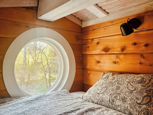 A bedroom with a round window, wooden walls, a bed with patterned bedding, and a hanging lamp.