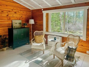 A cozy room with wooden walls, a green cabinet, two wicker chairs, a small table, and a window with a view of trees.