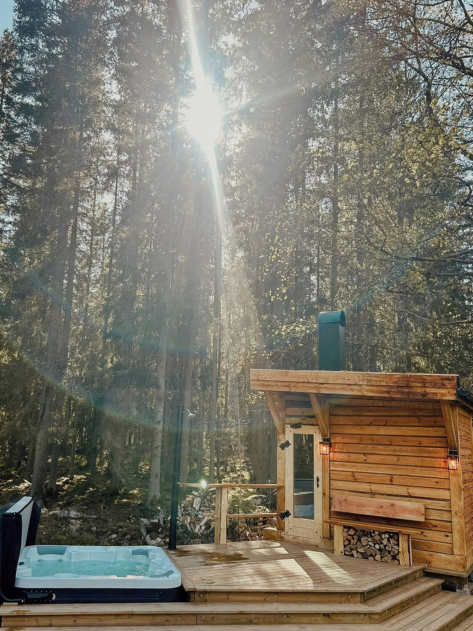 A wooden cabin with a hot tub and a deck, surrounded by a forest.