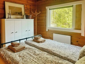 Een slaapkamer met een bed, twee kussens, een nachtkastje, een dressoir, een spiegel en een raam met uitzicht op bomen.