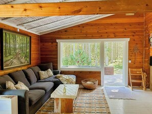 A cozy cabin interior with a wooden ceiling, a large window with a view of trees, a sofa with cushions, a small table, and a woven basket. - Log cabin with wood-fired sauna & Hot-Tub (Urshult)