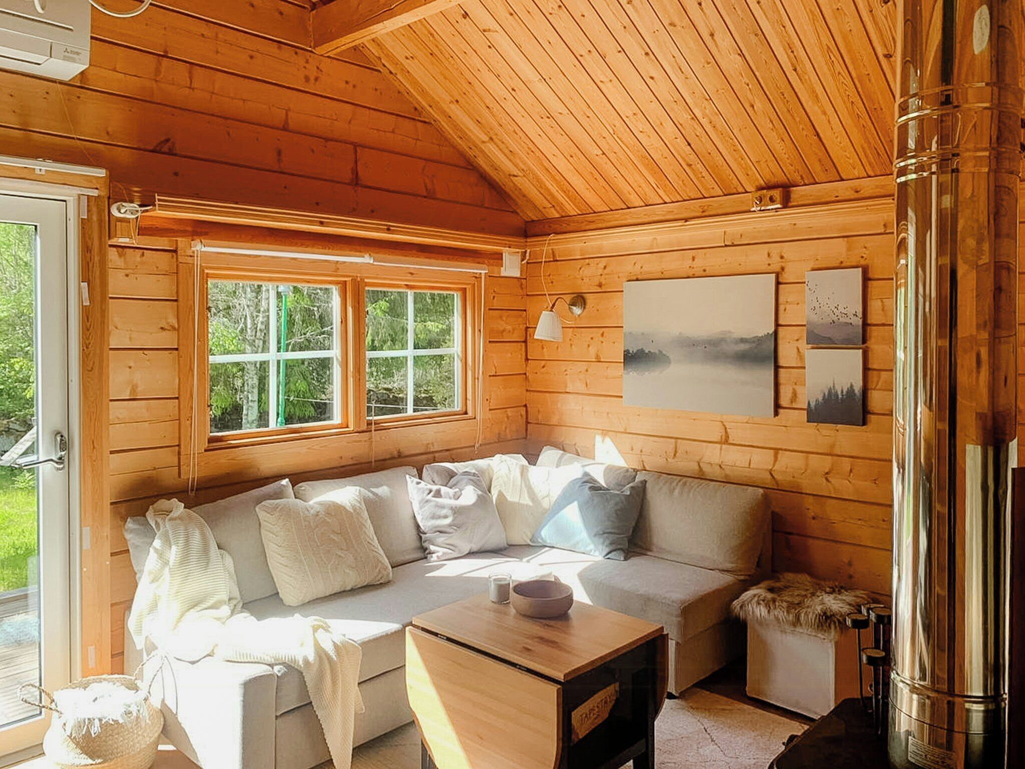 A cozy cabin interior with a wooden ceiling, a sofa with cushions, a table, and a window with a view of greenery.