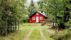 Ein rotes Haus mit weißem Tor und Fenstern, umgeben von Bäumen und einem grasbewachsen Weg, der davorführt.
